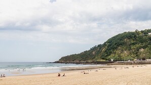 A beach with people relaxing and sunbathing, a rocky cliff with greenery, and a calm sea. - GRAN VIA SUITE: superior apartment for 6 PAX. 2min from the beach (San Sebastián)