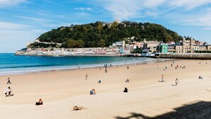 Una playa con gente tomando el sol y nadando, una colina con edificios, y un cielo despejado.