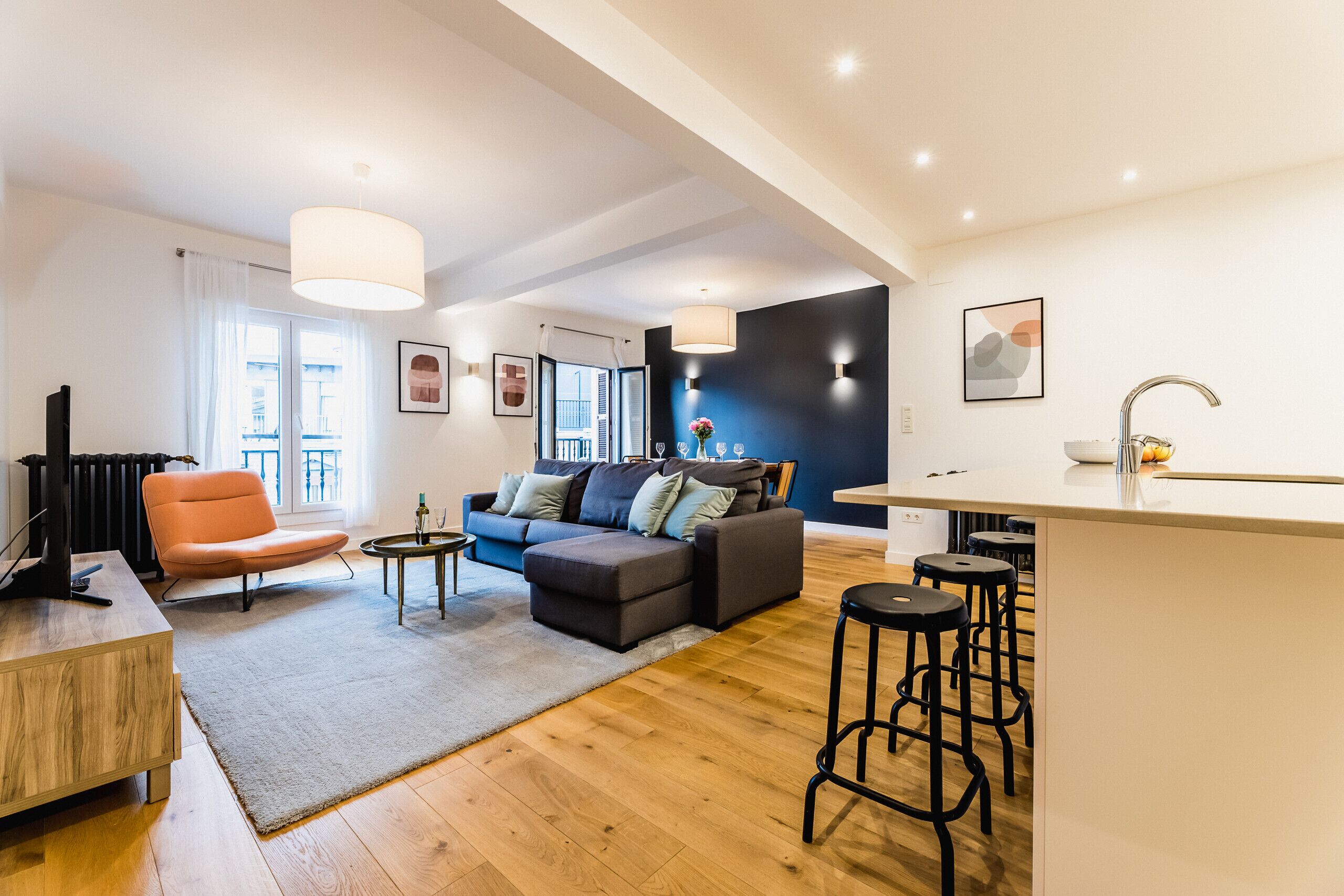 A modern living room with a sofa, ottoman, and a coffee table. There is a kitchen area with a bar stool and a dining table.