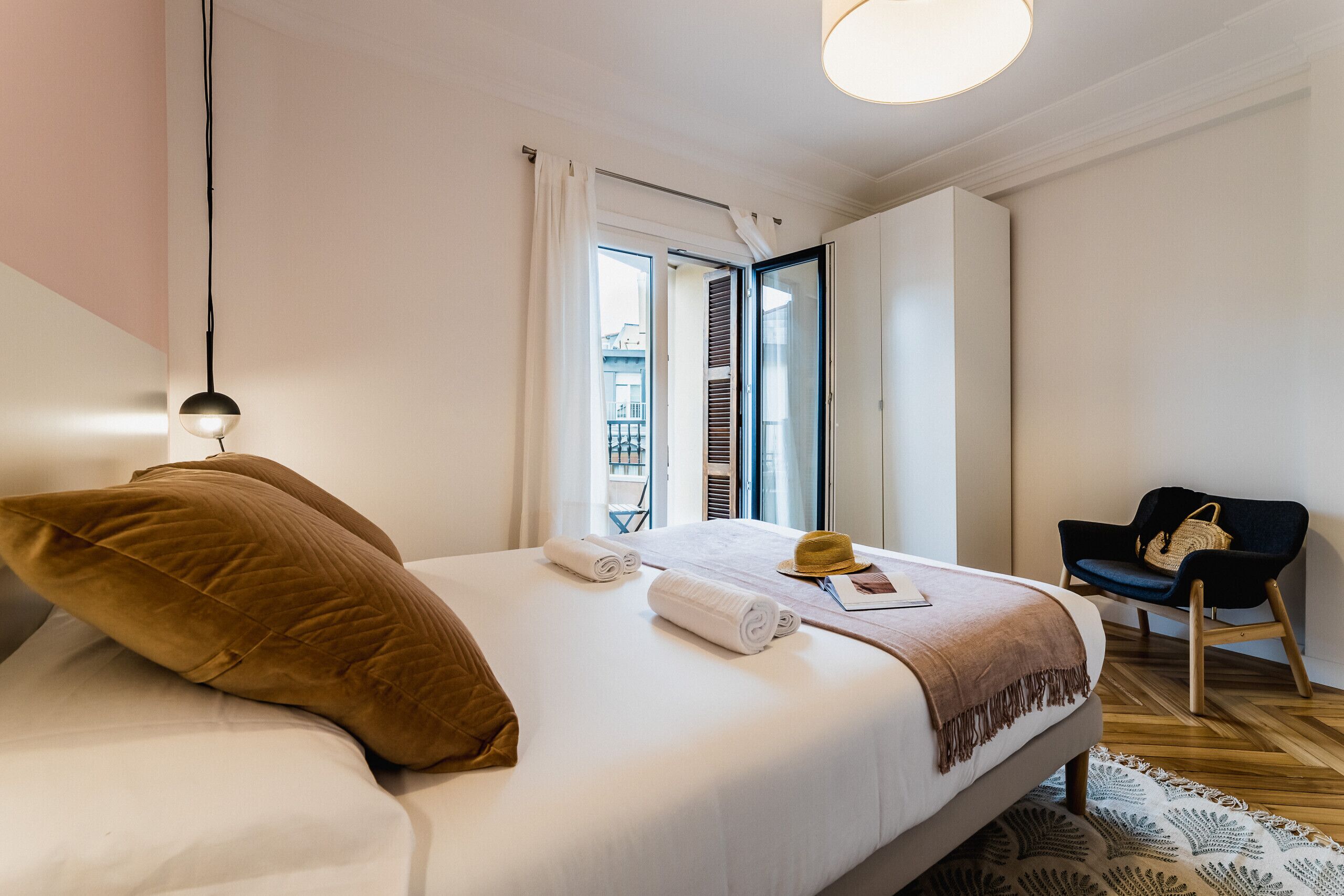 A neatly made bed with a brown pillow, a rolled towel, and a hat on the bed. There is a chair with a woven bag beside the bed.