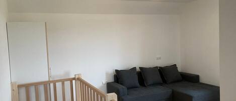Een moderne woonkamer met een houten trap, een donkergrijze hoekbank en één lamp in het plafond.