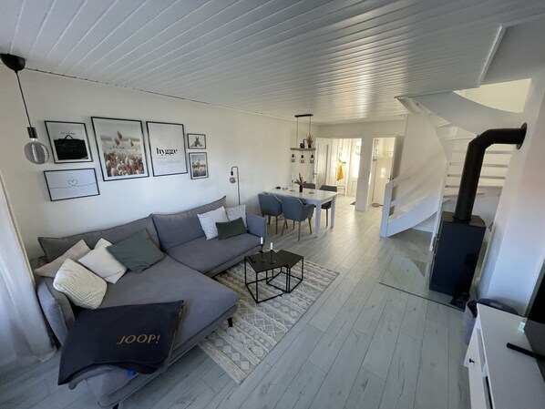 A modern living room with a grey sofa, a coffee table, and a dining area with chairs. There are framed pictures on the wall and a staircase leading to another level.