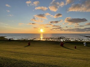 A grassy field with a sunset over the ocean, a pier, and a few chairs.