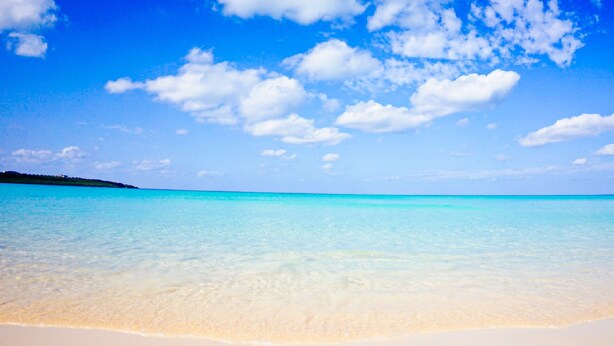 Una playa con aguas turquesas cristalinas y una orilla arenosa bajo un cielo azul con nubes dispersas.