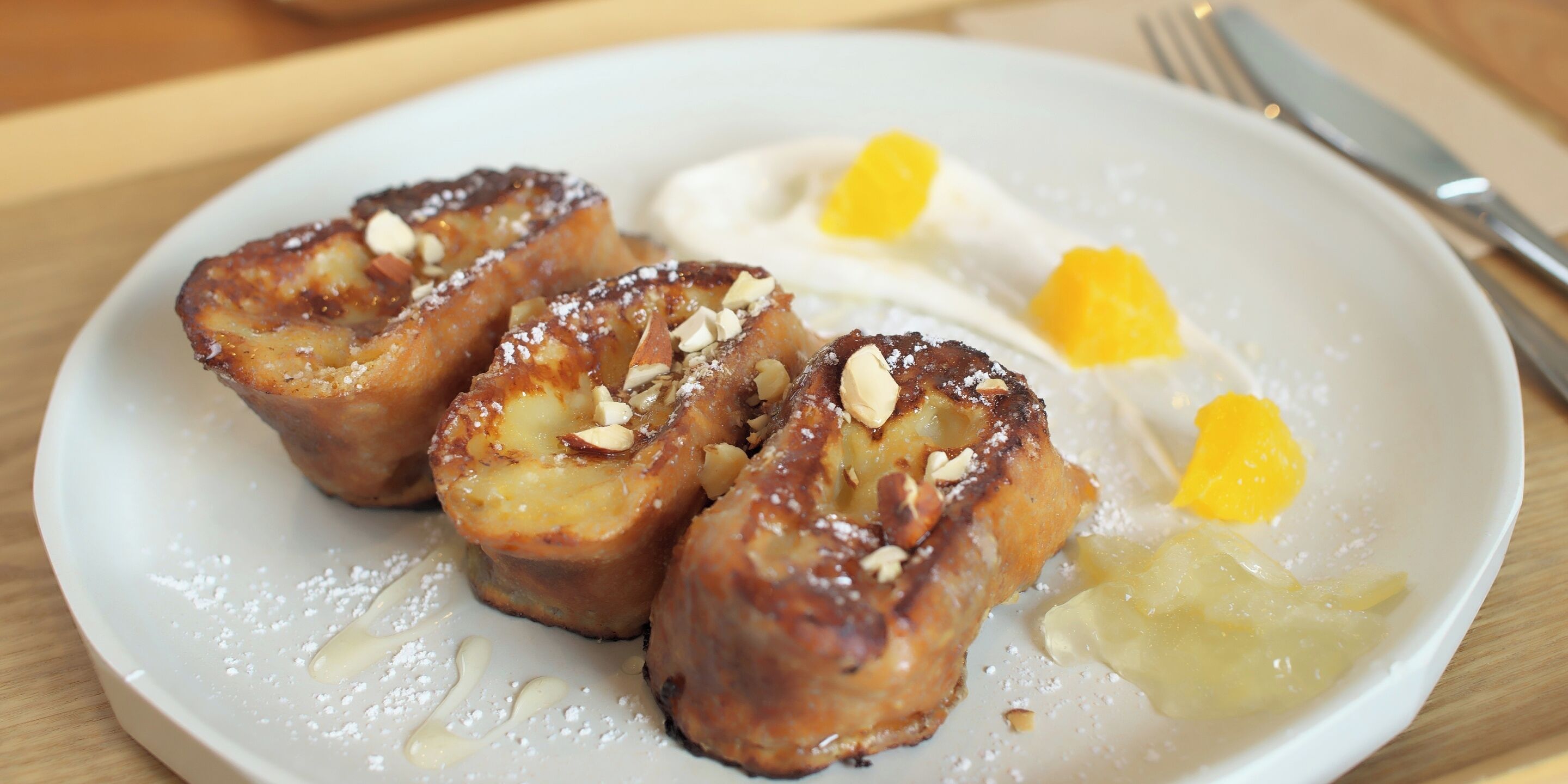 A plate with a dessert consisting of a baked pastry, possibly a tart, topped with sliced almonds and dusted with powdered sugar, accompanied by a dollop of cream and a slice of fruit.