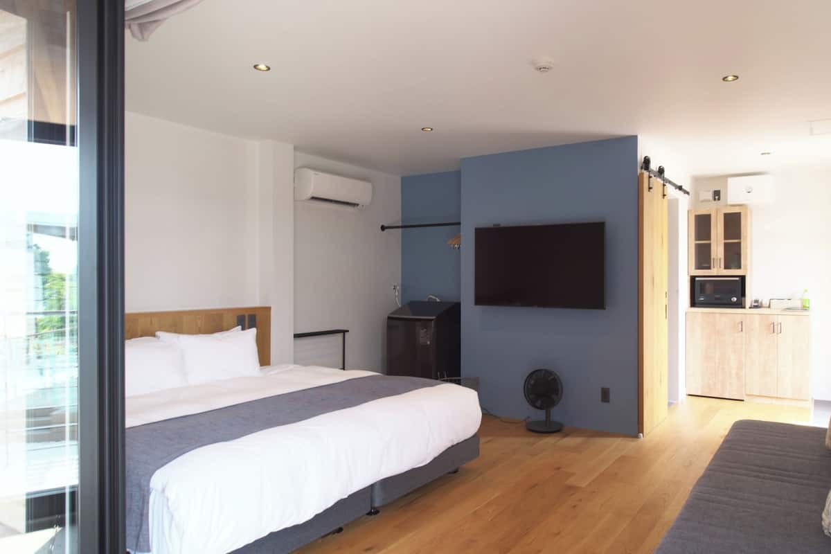 A modern bedroom with a large bed, a TV mounted on the wall, and a small kitchenette in the background.