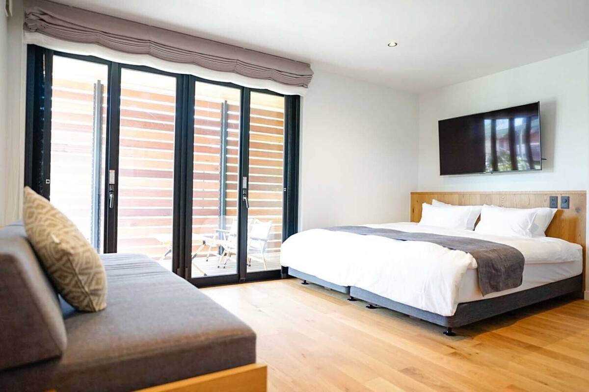 A bedroom with a bed, a sofa, a TV, and a sliding glass door to the outside.