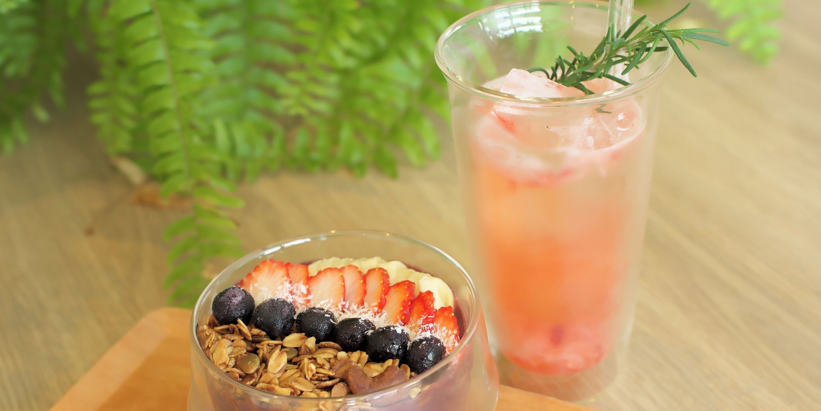 Um copo de bebida gelada com um ramo de alecrim, um prato de granola com frutas, e uma mesa de madeira com uma planta verde ao fundo.