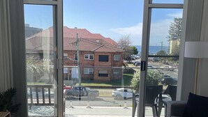 Een kamer met een grote glazen schuifdeur met uitzicht op de straat, een grijze bank, een wit voetenbankje en een kleine plant.