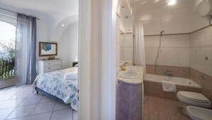 Une chambre à coucher avec un lit, une table de chevet et une fenêtre avec des rideaux. L’image montre également une salle de bain avec une baignoire, des toilettes et un lavabo.