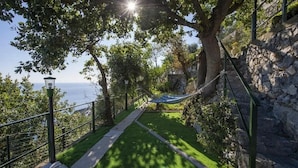 Un sentier mène à un hamac suspendu sous un arbre, avec un mur de pierre d’un côté et une vue sur la mer de l’autre.