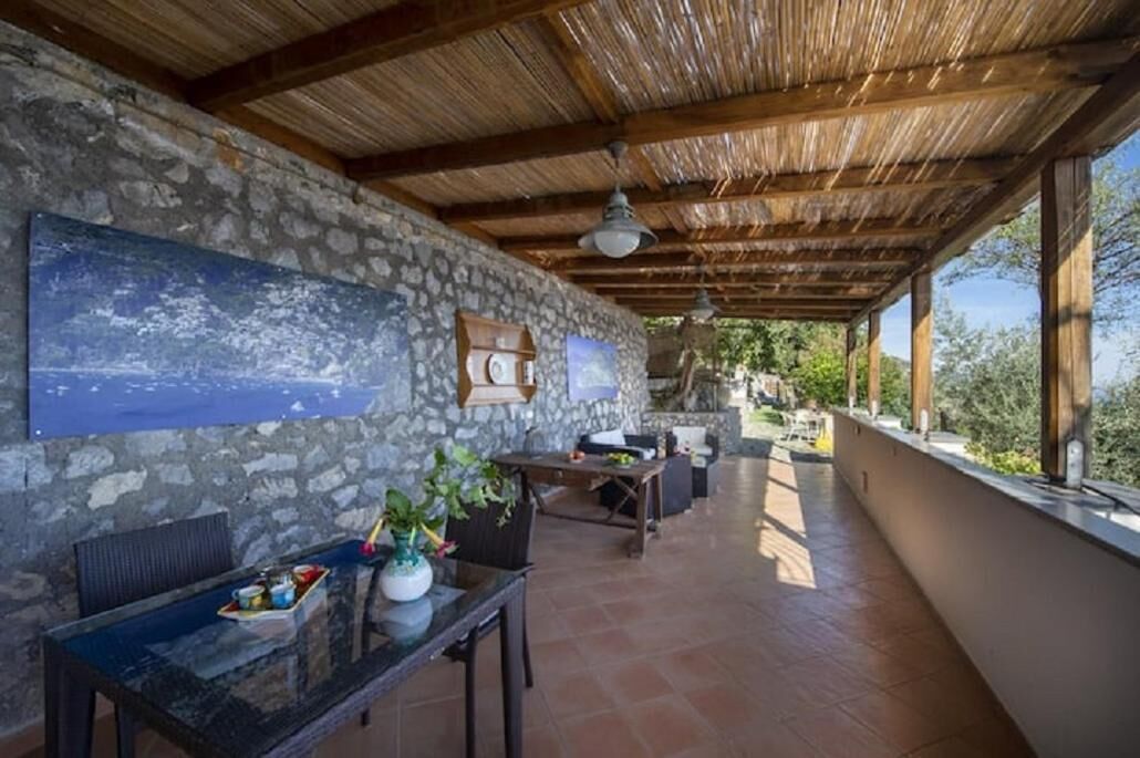 Un espacio al aire libre techado con paredes de piedra, techo de madera y una mesa de comedor con sillas.