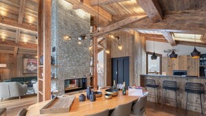 A rustic interior with a stone fireplace, wooden beams, a dining area with a long table and chairs, and a bar area with stools.