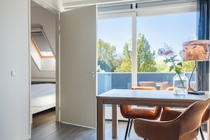 Ein Zimmer mit einem Bett, einem Esstisch mit Stühlen, einer Lampe und Blick auf Bäume durch ein Fenster.