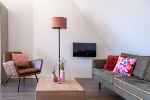 A modern living room with a brown leather armchair, a grey sofa, a wooden coffee table, a tall floor lamp, and a flat-screen TV mounted on the wall.