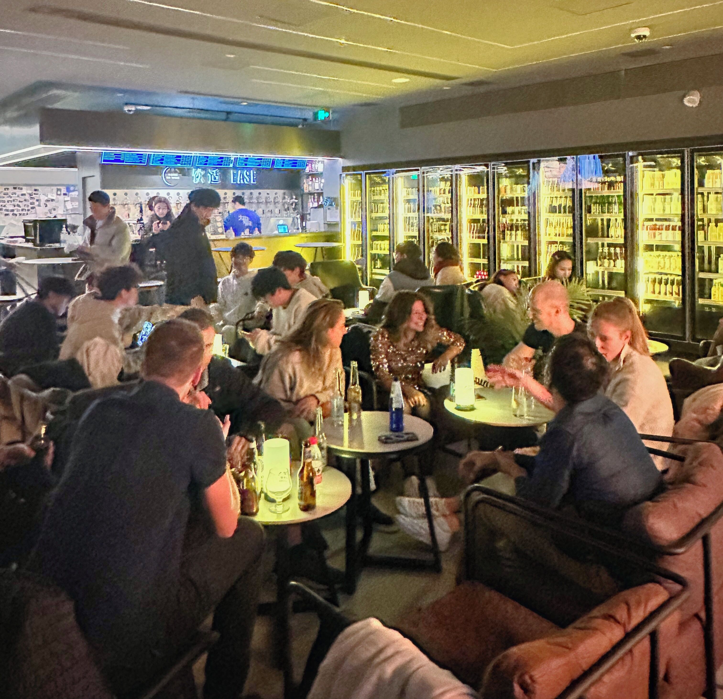 A crowded bar with patrons seated at tables, a bartender at the counter, and shelves stocked with bottles behind the bar.