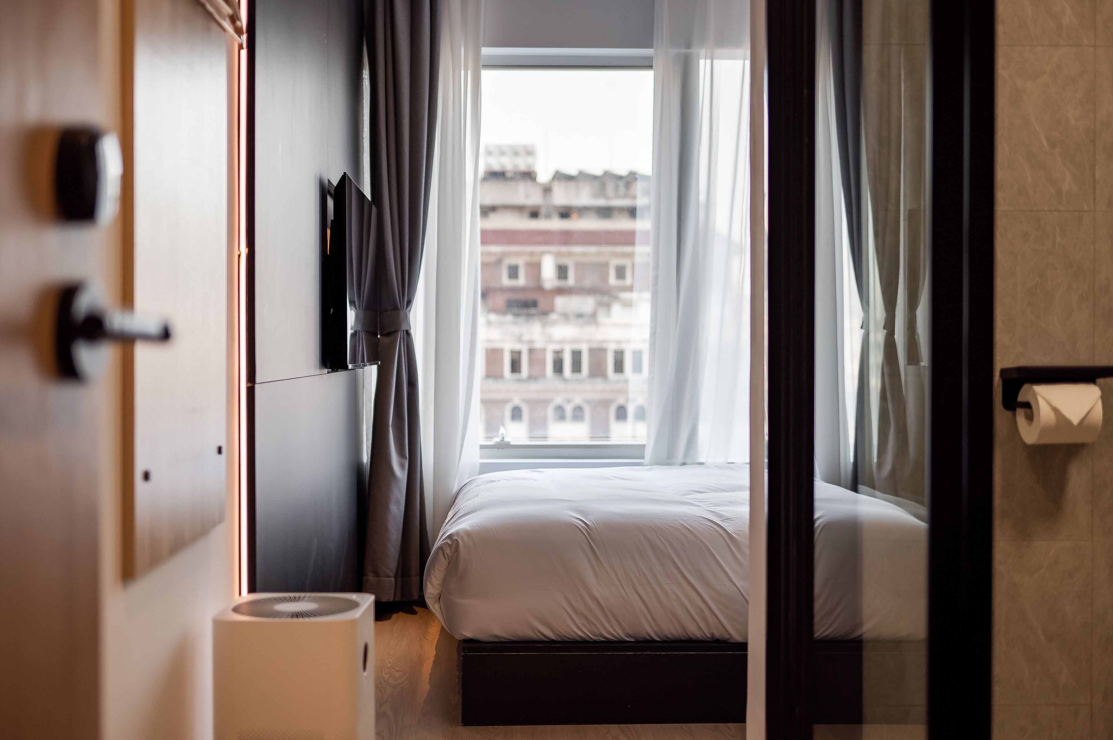 Habitación de hotel con cama, ventana con cortinas, vistas a edificios y un televisor montado en la pared.