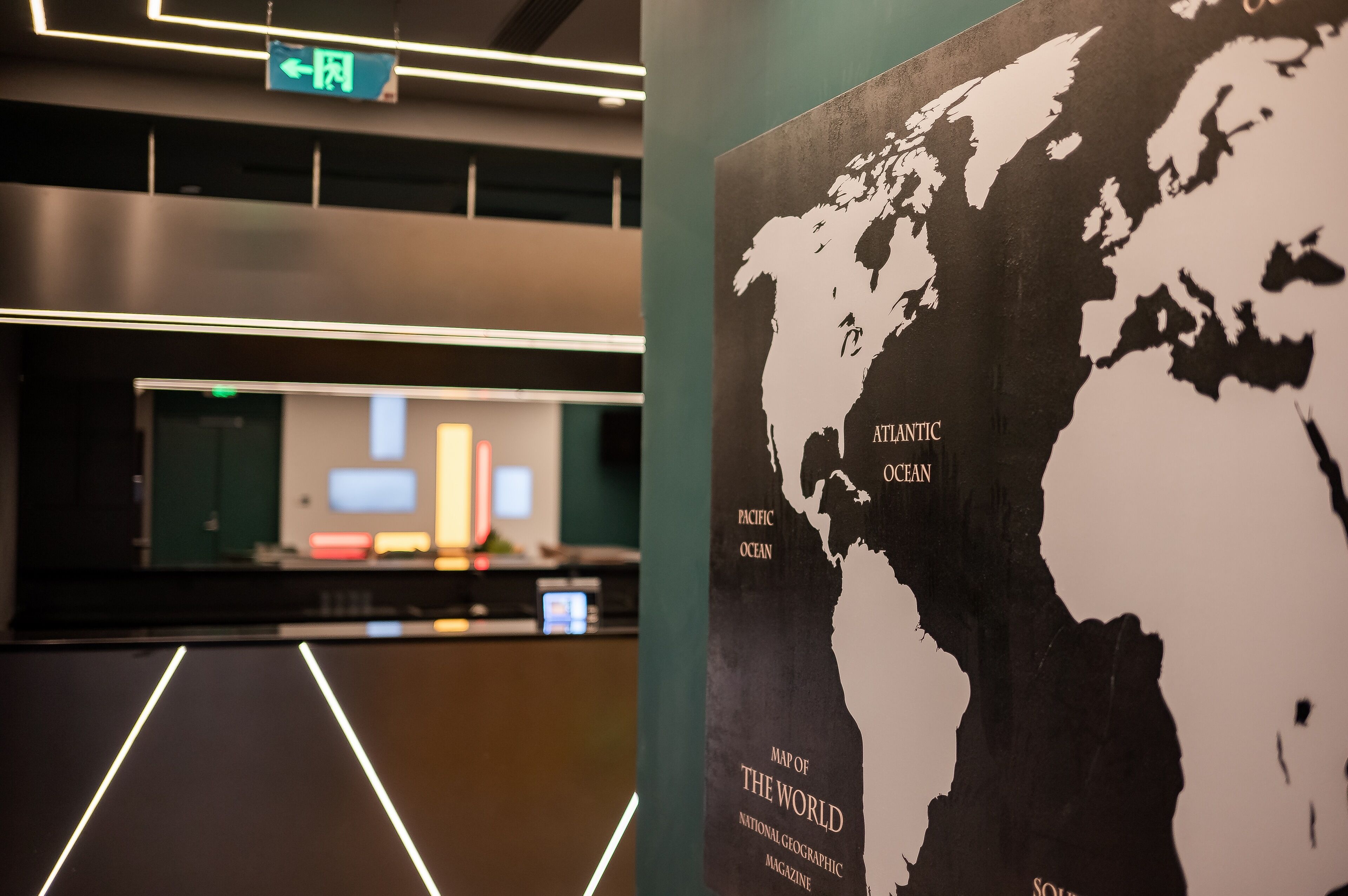 Un mostrador de recepción con un monitor de computadora, un letrero de salida verde y un cartel con un mapa del mundo.