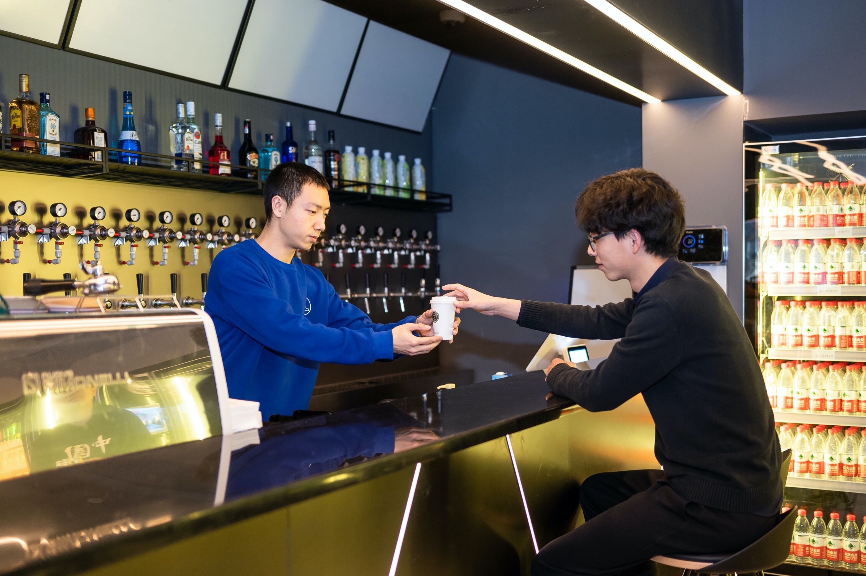 Un espace bar avec une variété de bouteilles d’alcool exposées sur des étagères et un présentoir réfrigéré contenant des bouteilles d’eau.