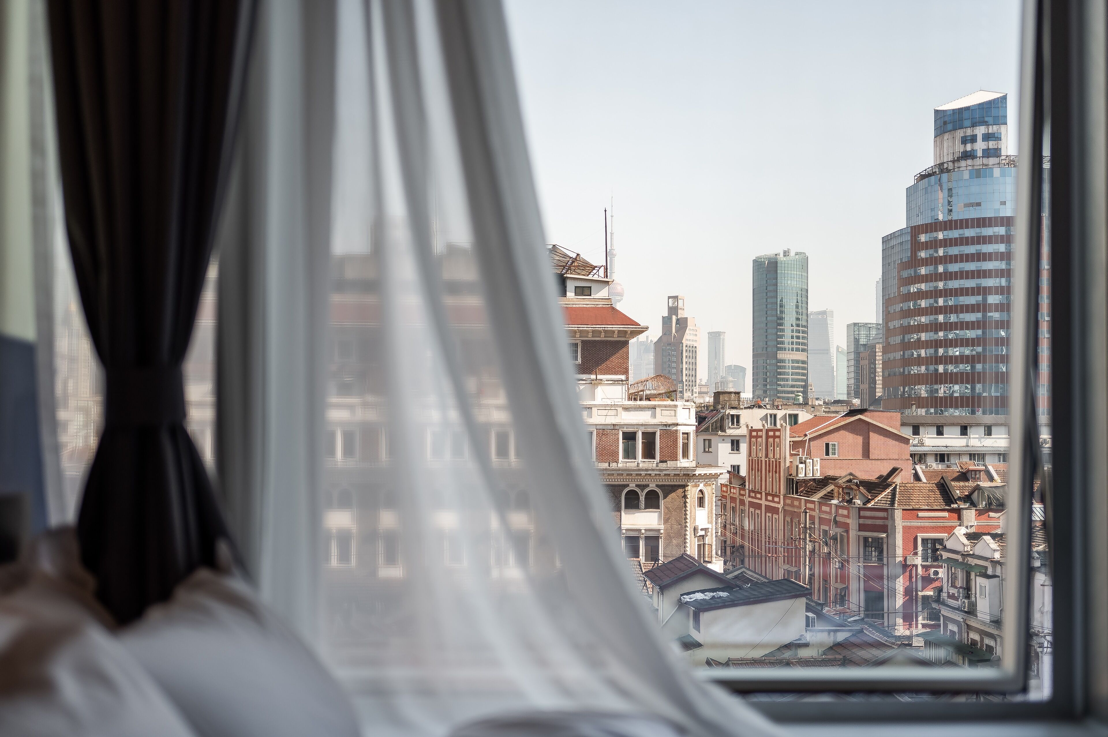 Blick aus einem Fenster mit transparenten Vorhängen auf eine Stadtlandschaft mit modernen und historischen Gebäuden.