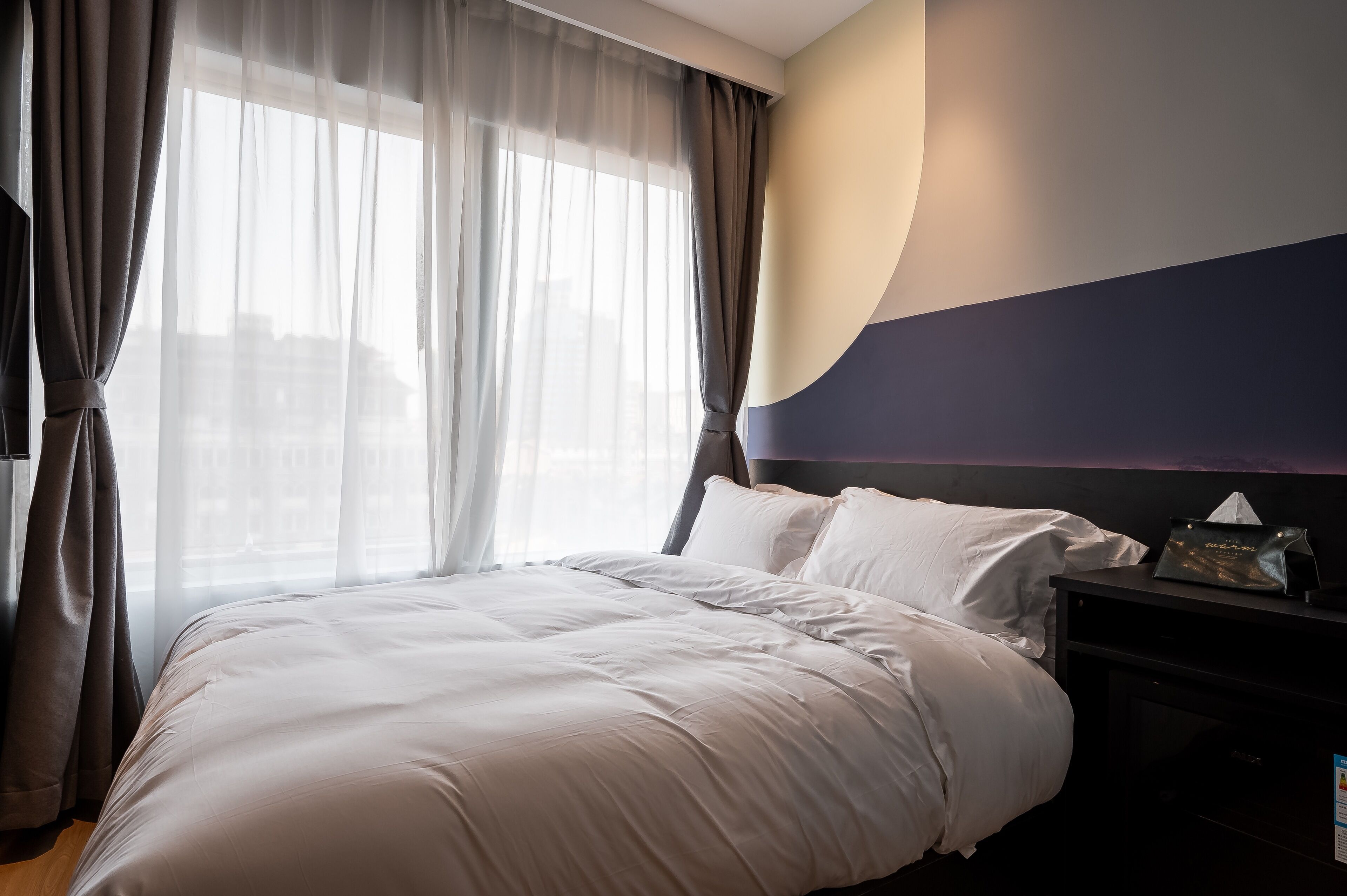 A hotel room with a large bed, a nightstand, and a window with sheer curtains.