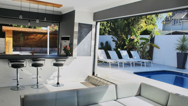 A modern outdoor living area with a pool, bar stools, and lounge chairs.
