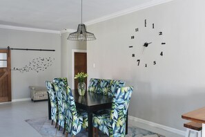 Un coin repas avec une table en bois, des chaises ornées de motifs de feuilles tropicales, une horloge murale et un luminaire suspendu.