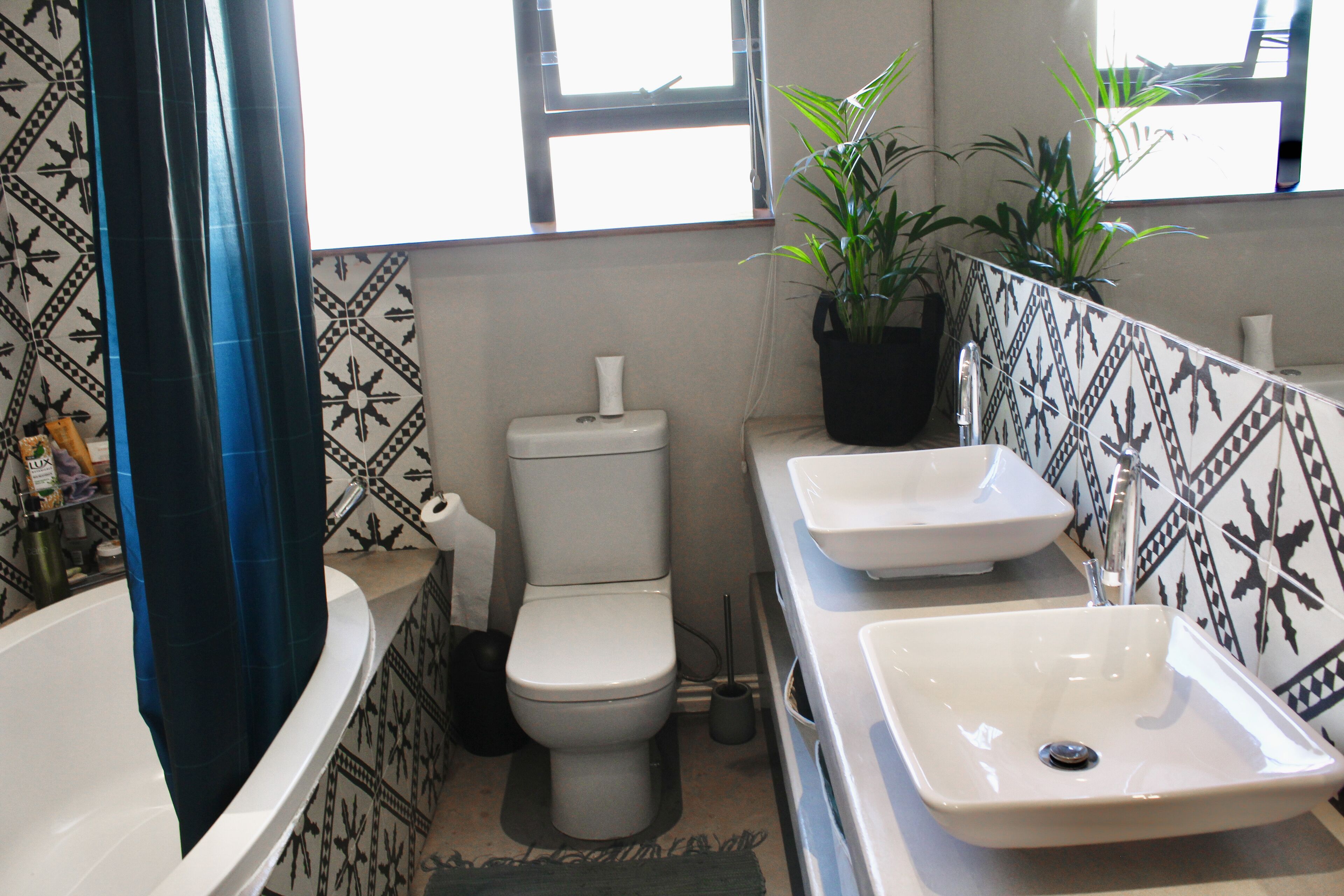 Ein Badezimmer mit einer weißen Badewanne, einer Toilette und zwei Waschbecken. Die Wände sind mit dekorativen Fliesen versehen und über der Toilette befindet sich ein Fenster.