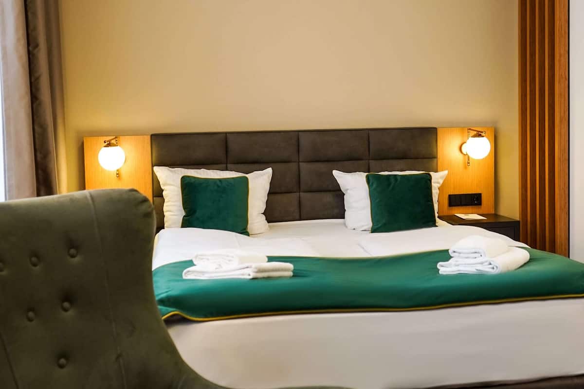 Een hotelkamer met een groot bed, twee bedlampen, een groene beklede fauteuil en een kamerbreed tapijt met dessin.