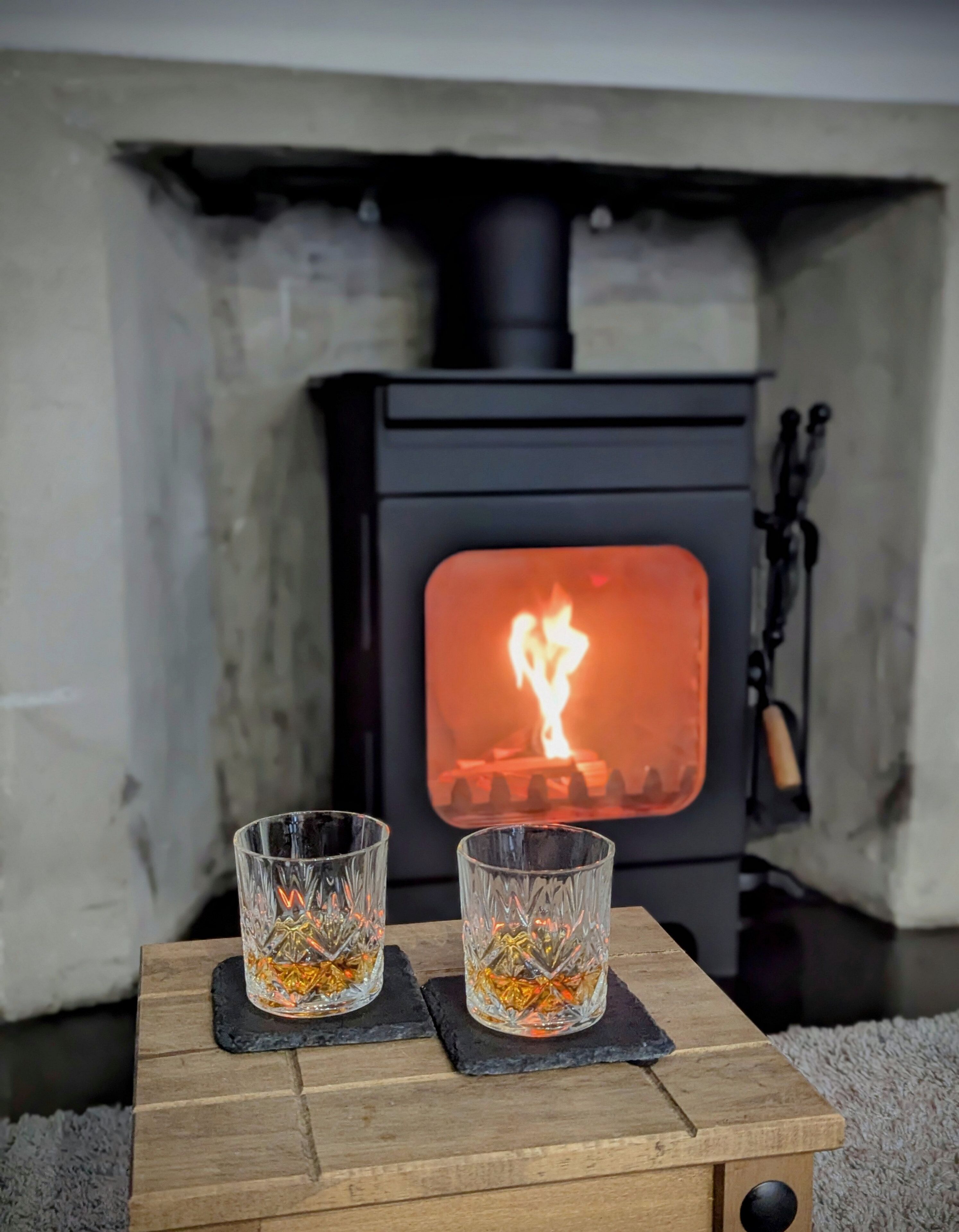 Una estufa de leña con dos vasos de una bebida dorada sobre una pequeña mesa de madera.