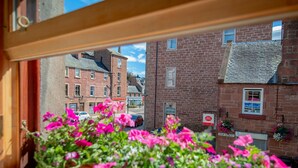 A view through a window with pink flowers in the foreground, a brick building with a 'POST OFFICE' sign, and a street with parked cars. - Unique 2 Bedroom Flat Kirrie Scotland (Kirriemuir)