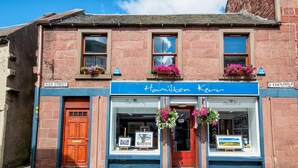 A building with a blue storefront labeled "Hamilton Kerr" and a wooden door on the left. - Unique 2 Bedroom Flat Kirrie Scotland (Kirriemuir)