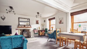 A cozy living room with a blue sofa, wooden dining table, and chairs. There is a fireplace, a television, and a large mirror on the wall. - Unique 2 Bedroom Flat Kirrie Scotland (Kirriemuir)