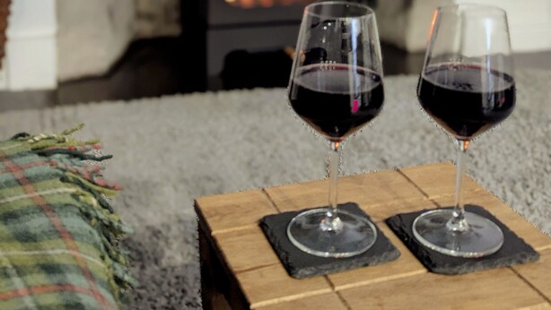 A cozy living room with a fireplace, two glasses of red wine on a wooden table, and a plaid throw blanket on a couch. - Unique 2 Bedroom Flat Kirrie Scotland (Kirriemuir)