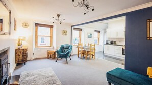 A cozy living room with a blue armchair, a small table, and a fireplace. There is a kitchen visible through an open doorway. - Unique 2 Bedroom Flat Kirrie Scotland (Kirriemuir)