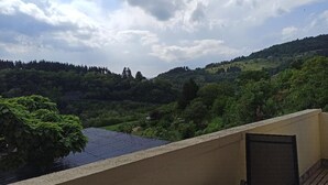 A balcony with a table and chairs, overlooking a lush green landscape with hills and trees under a partly cloudy sky. - Logement Calme et Spacieux Avec vue à 400 M D'un Plan D'eau (Lamastre)