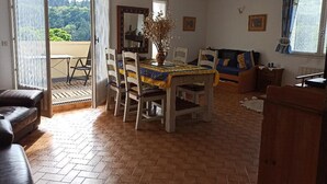 A dining area with a table set for two, a sofa, and a view of greenery outside. - Logement Calme et Spacieux Avec vue à 400 M D'un Plan D'eau (Lamastre)