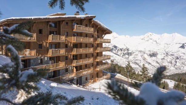 Un edificio de madera de varios pisos con balcones, rodeado de montañas nevadas y pinos.