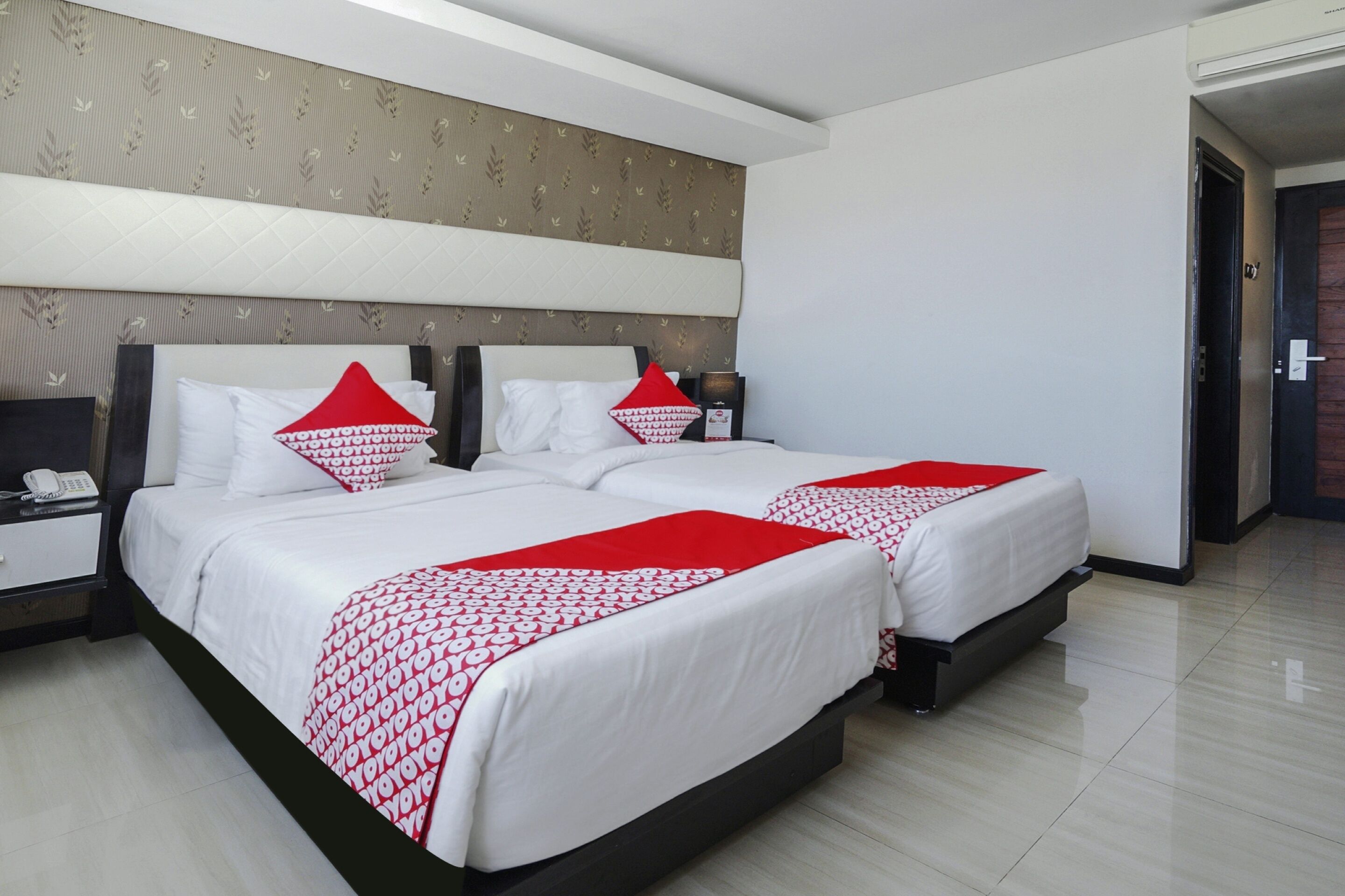 A hotel room with two beds, each with white bedding and red accents, a bedside table with a phone, and a nightstand with a lamp.
