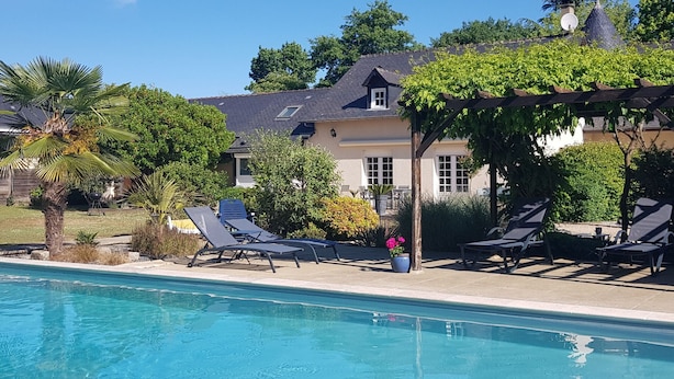 Une piscine avec des chaises longues et une pergola, entourée de verdure, avec une maison en arrière-plan.