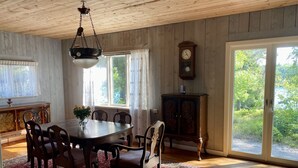 A dining room with a wooden table, chairs, and a red patterned rug. There is a large window with curtains, a wooden cabinet, and a wall-mounted clock. - Cottage with fantastic lake view!  (vittsjö)