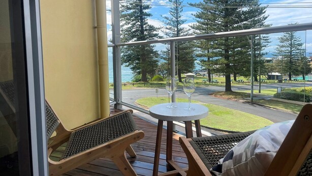 Un balcón con vista a un parque y un lago. Hay dos sillones de mimbre y una pequeña mesa redonda con dos copas de vino.
