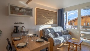 A cozy room with a wooden dining table set for four, a sofa with patterned cushions, and a view of snowy buildings through large windows. - Renovated apartment near slopes with wifi (Val Thorens)