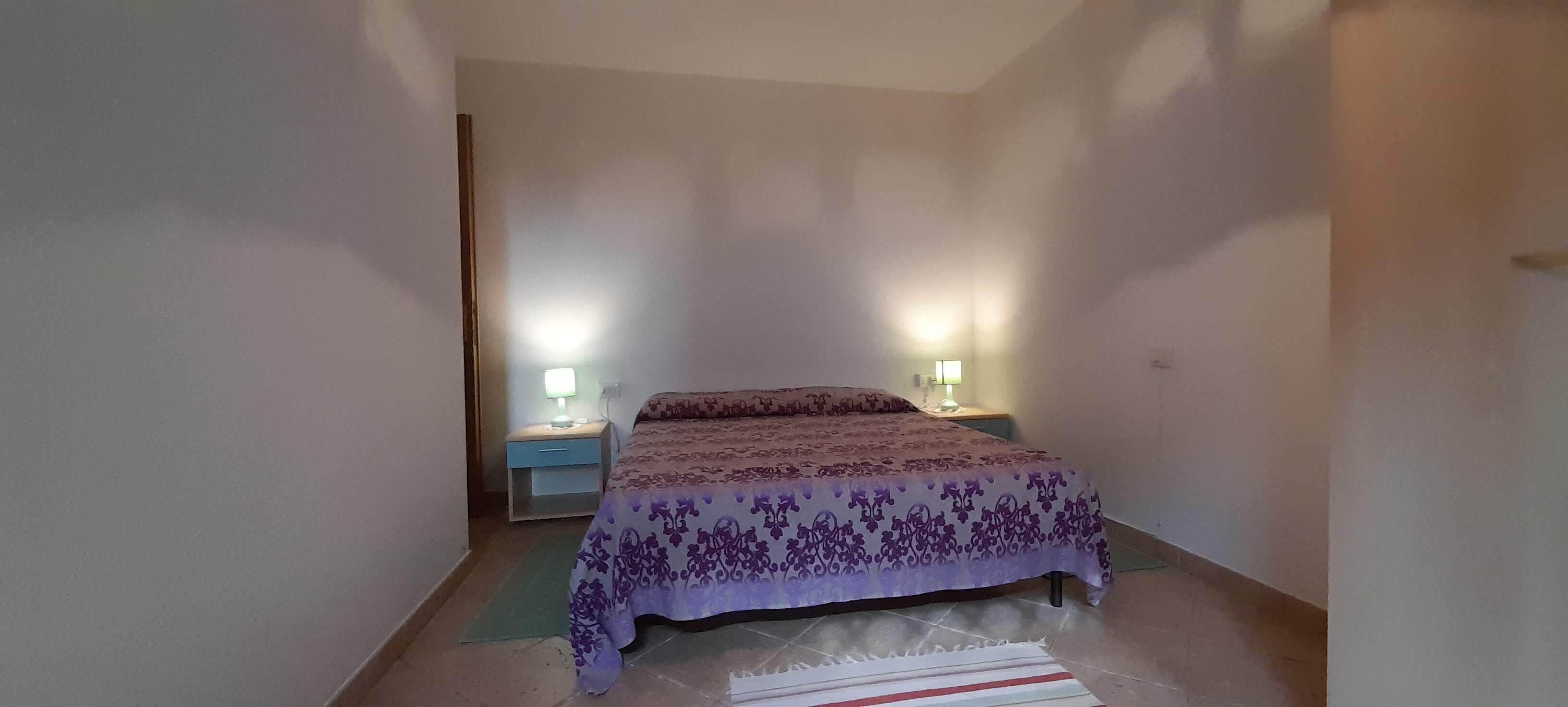 A small, narrow room with a bed, two bedside tables with lamps, and a plain wall.
