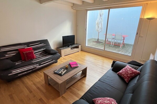 Una sala de estar moderna con un sofá negro, una mesa de centro de madera y un televisor sobre un pequeño soporte. Hay un gran ventanal corredero que da a un área exterior con una mesa y sillas.