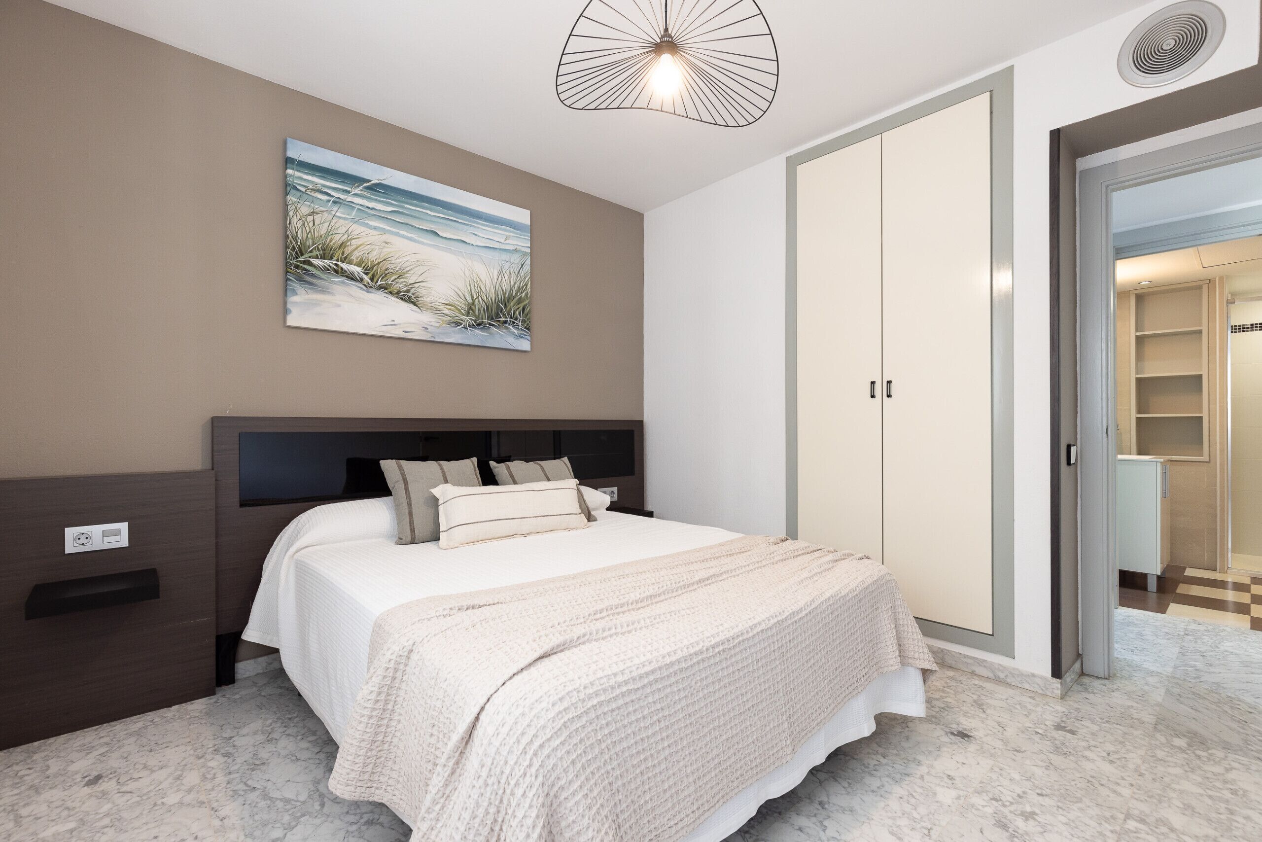 Un dormitorio con cama, mesita de noche, un cuadro, armario y baño visible a través de una puerta abierta.