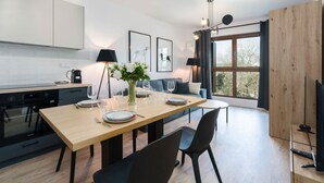 A modern kitchen with white cabinets, a dining table set for four, and a large window with a view of trees. - Comfortable Apartment | Balcony, Parking, next to the Fair (Poznan)