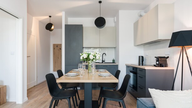 A modern kitchen with a central island, dark cabinetry, and a dining area with a wooden table and black chairs. - Comfortable Apartment | Balcony, Parking, next to the Fair (Poznan)