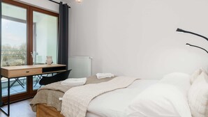 A bedroom with a bed, a desk, and a chair. There is a window with curtains, a radiator, and a hanging light fixture. - Comfortable Apartment | Balcony, Parking, next to the Fair (Poznan)