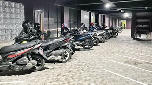 Fileira de motocicletas estacionadas em área coberta, com piso de azulejos e parede de vidro em um dos lados.